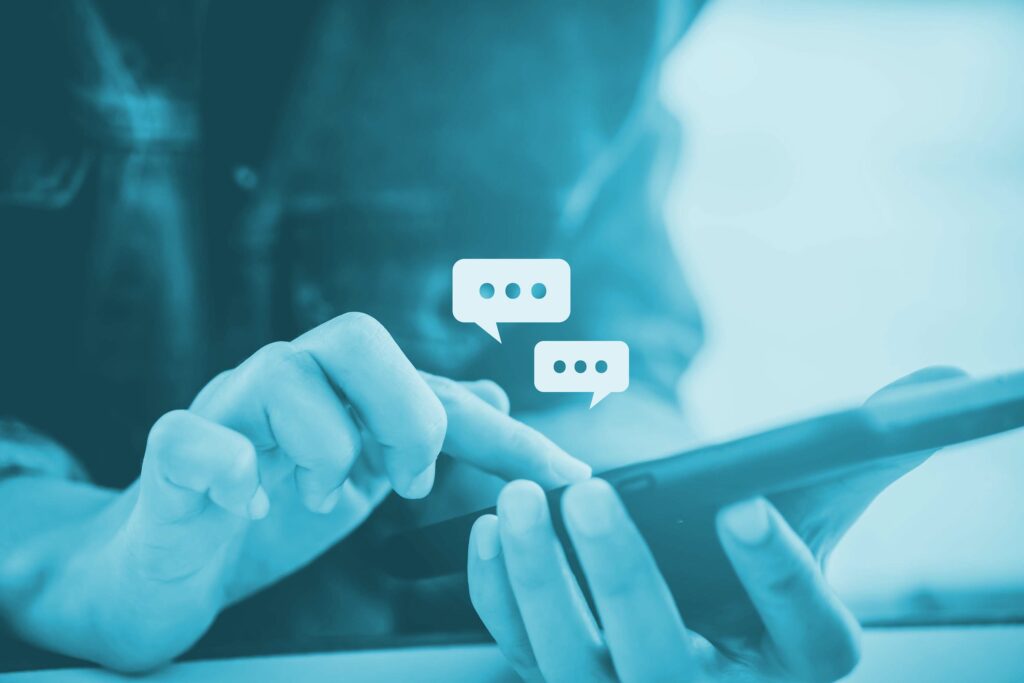 Main tenant un smartphone en gros plan, avec des icônes de bulles de dialogue illustrant une conversation ou une messagerie instantanée, sur un filtre bleu.